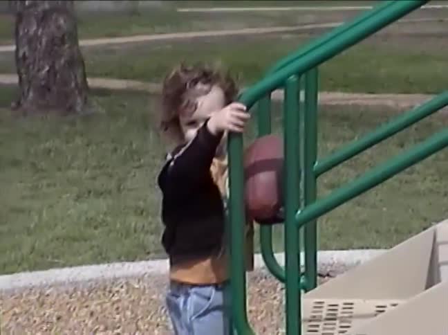 A determined toddler with curly hair struggles but eventually climbs the playground stairs while holding onto a football