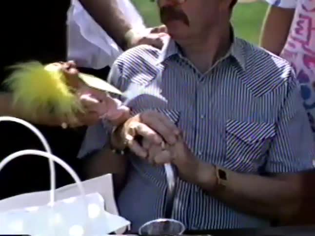 A man with a mustache and cowboy hat is presented with a troll doll as a gift at a sunny outdoor gathering