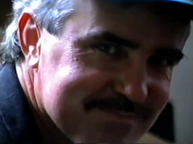 Candid home video zooms in on a smiling man with a mustache and cap, then pans to an older man in a doorway