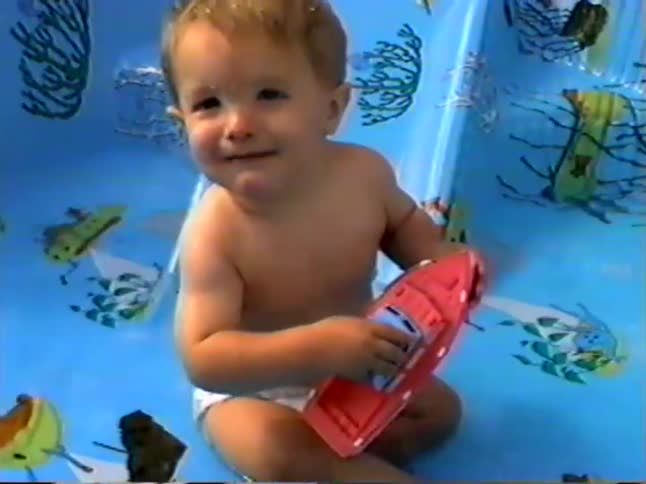 An adorable baby boy sits in a small kiddie pool while playing with his red toy boat