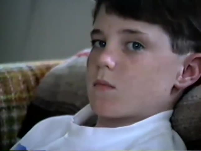 A young boy with blue eyes looks seriously at the camera before suddenly opening his mouth wide in a shocked expression in this vintage home video