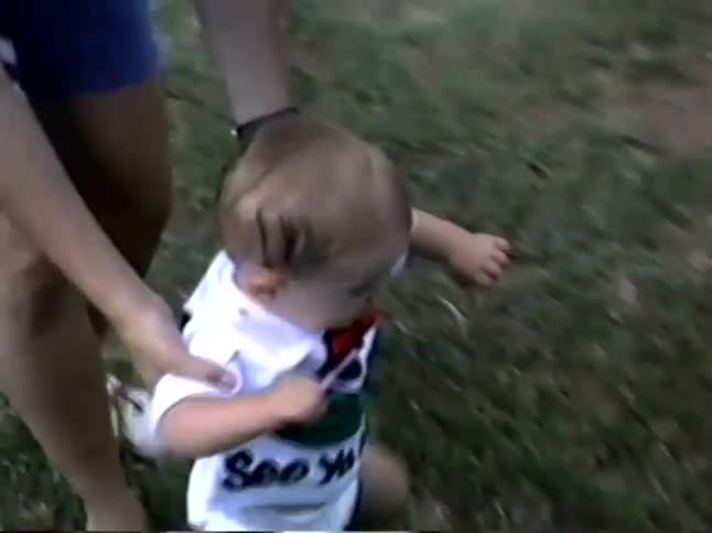 Vintage home video footage shows a parent helping their baby take their first wobbly steps on a grassy lawn