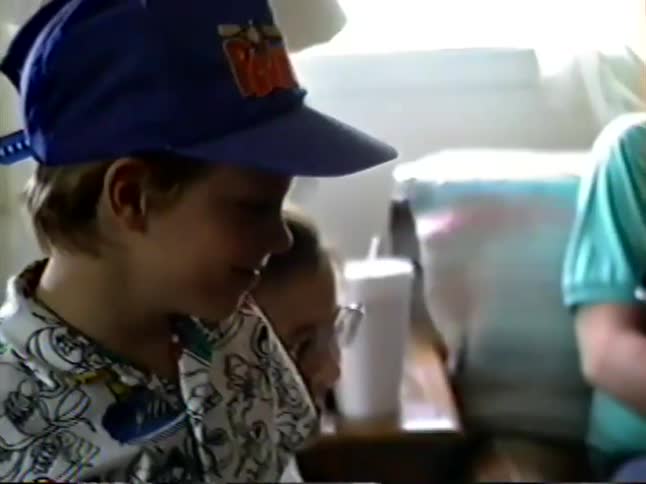 A sweet vintage VHS home movie from the 1990s shows a young boy in a baseball cap smiling before the camera pans to his family sitting on a couch