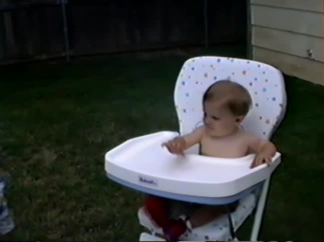 A cute baby in a high chair looks fussy until siblings bring a slice of birthday cake in this vintage home video