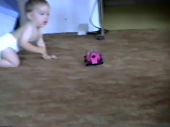 A cute baby crawls on the floor and curiously watches a pink remote-controlled car in a vintage home video