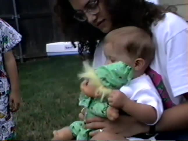 A sweet toddler sits on a woman's lap outdoors and playfully interacts with a new troll doll toy