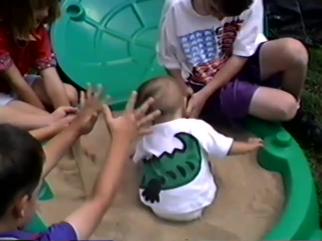 A group of older children play together in the sand with a baby sitting in a green turtle-shaped sandbox