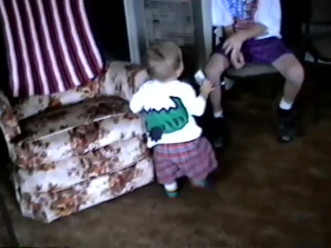 A vintage home video shows a toddler in plaid pants taking wobbly steps in a 1990s living room as their older brother watches