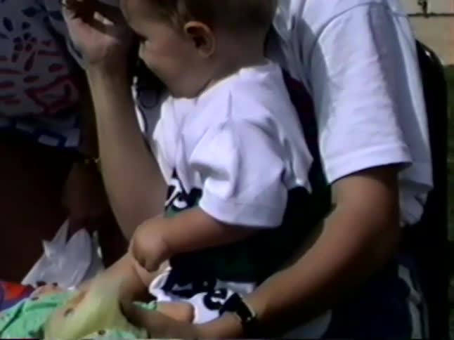 Vintage VHS footage shows a curious baby boy sitting on his mother's lap and playing with a ball and a troll doll outdoors on a sunny day