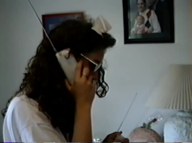 A young mother talks on a vintage cordless phone while her toddler son stands nearby in their 1990s living room