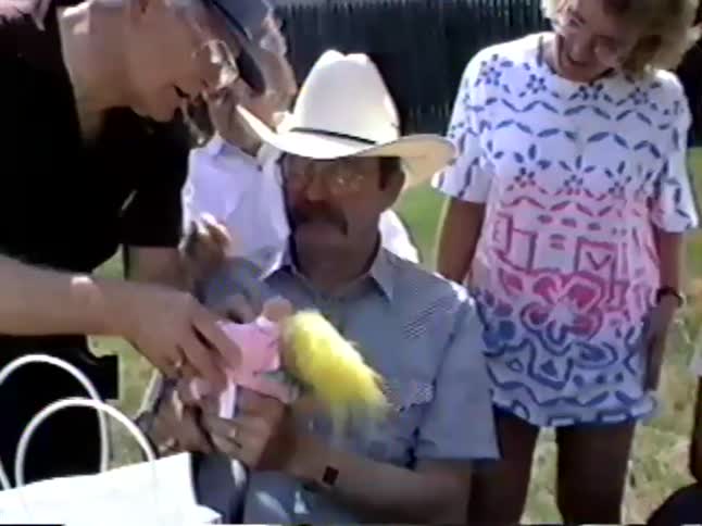 A man in a cowboy hat is surrounded by family as he receives a funny troll doll as a gift at an outdoor party in this vintage home video