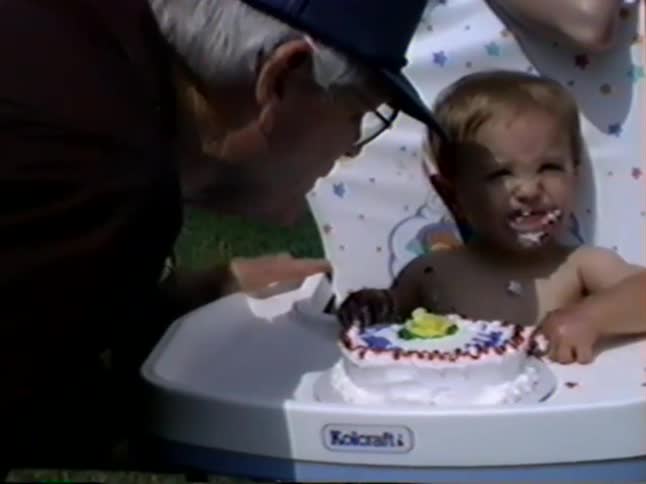 Vintage footage from the 1990s shows a baby joyfully eating and smashing their birthday cake in a high chair while family watches