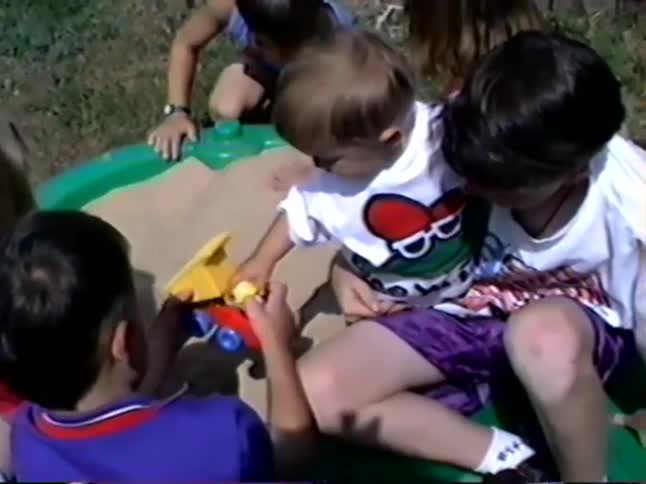 A group of young children play together with toys in a green sandbox on a sunny day in a backyard