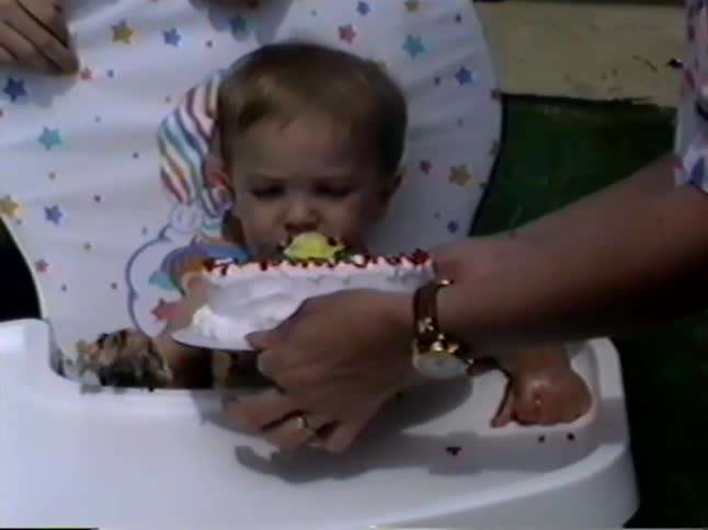 A baby in a high chair gleefully smashes and eats their first birthday cake while their family watches in a vintage home video
