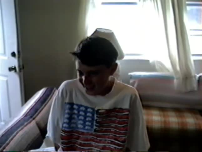 A vintage home video shows a young boy with a black eye smiling shyly for the camera in his bedroom