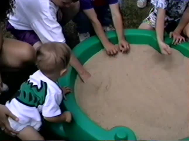An adult helps a toddler get into a green turtle-shaped sandbox where his initial curiosity turns to tears