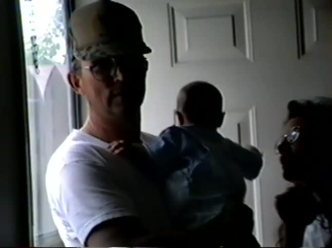 A candid vintage home video captures a family gathering with a grandfather holding a baby in silhouette by a bright doorway