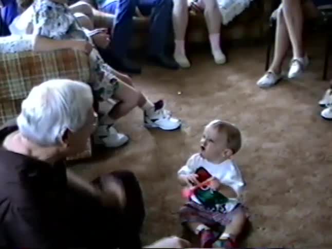 An endearing moment between a grandfather and his grandchild playing with a party noisemaker in a living room full of family