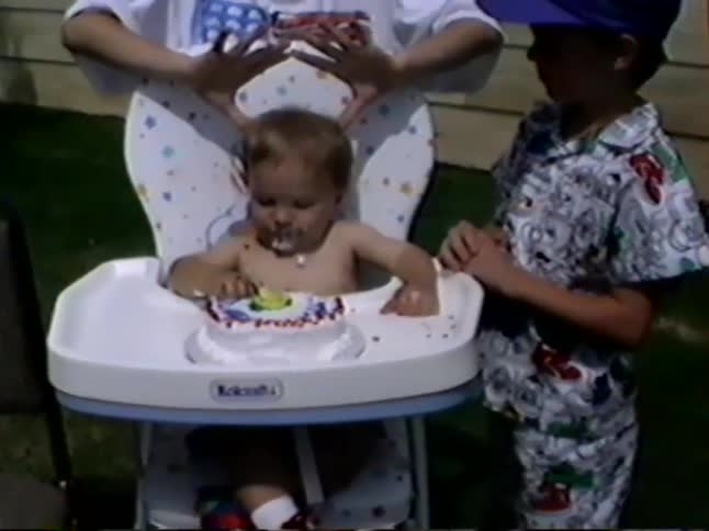 A shirtless baby in a highchair enjoys a messy first encounter with a birthday cake during a backyard party