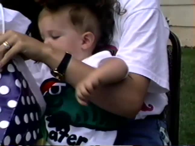 A mother holds her baby on her lap as they open a gift from a polka-dotted bag in the backyard during a celebration