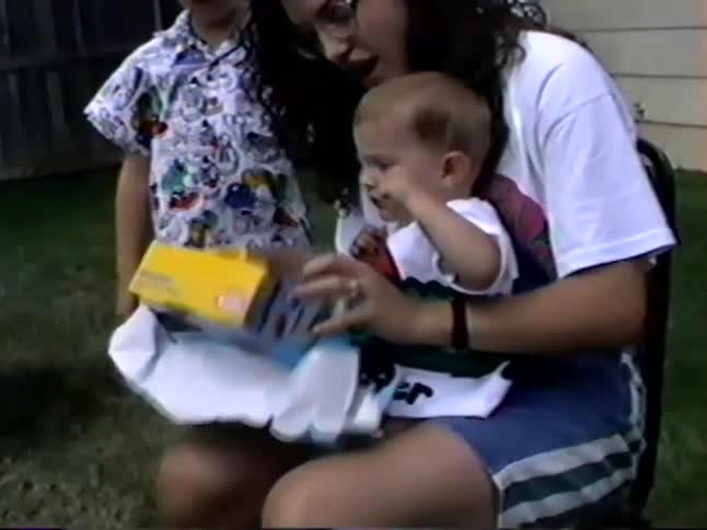 A mother helps her baby boy unwrap a birthday gift in the backyard, revealing a colorful toy dump truck in this vintage home video
