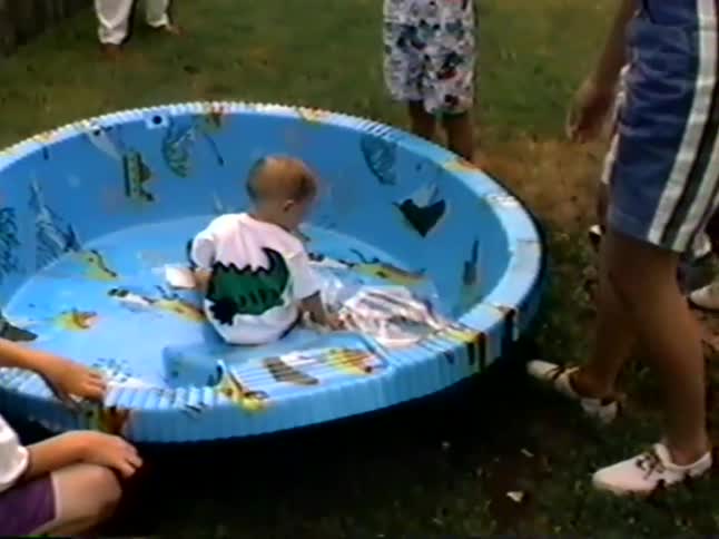 A mother carefully places her baby into an empty blue wading pool in a grassy backyard during a family gathering