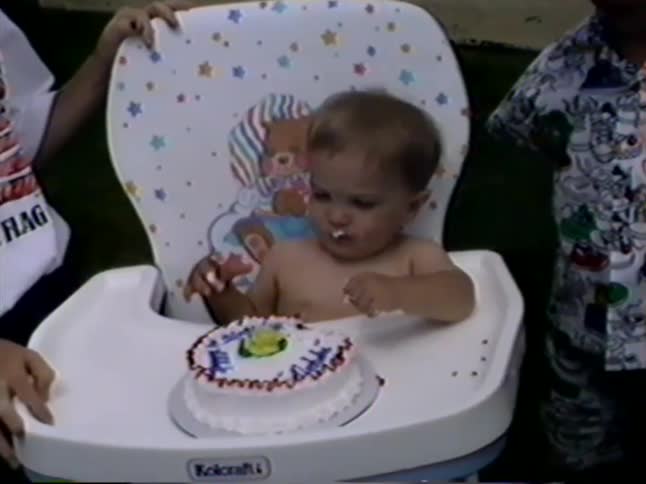 A toddler sitting in a high chair makes a mess while happily eating their birthday cake with their hands