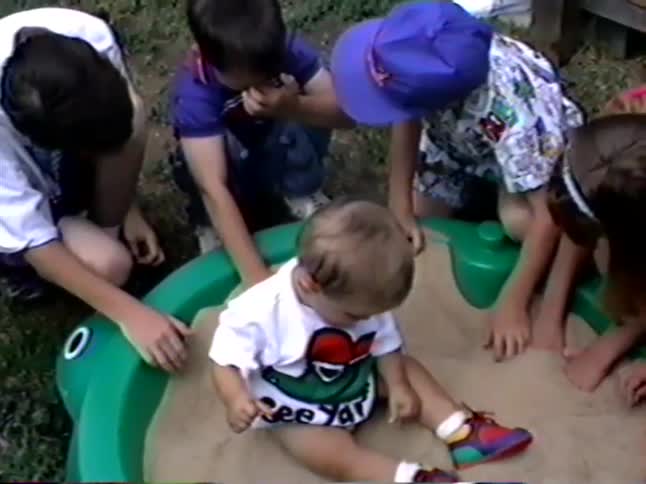 A group of kids from the 90s play in a green turtle sandbox with a cute toddler sitting in the middle