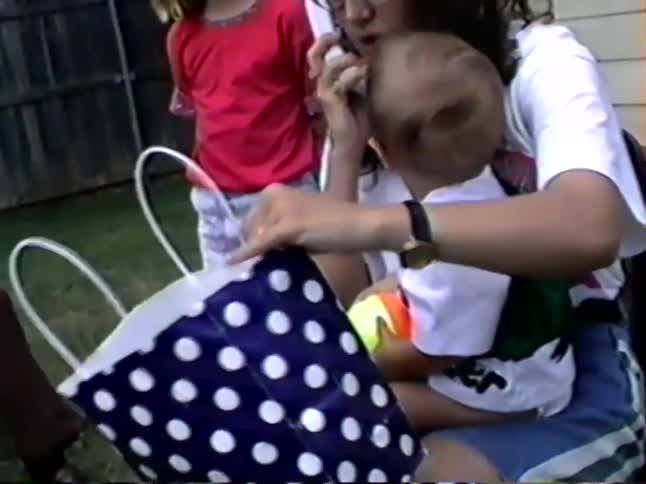 A vintage home video shows a mother holding her baby at an outdoor party as they open presents and she answers a phone call