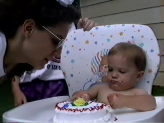 A cute baby boy in a high chair messily eats his first birthday cake with his hands and shares a bite with his mom