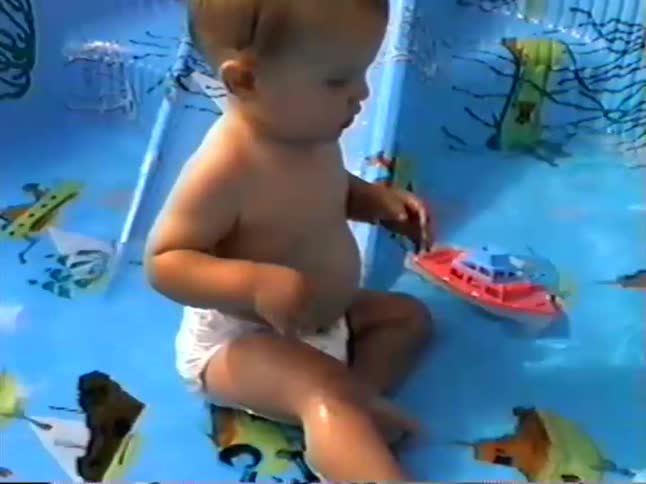 A cute baby sits in a small kiddie pool, playing with a toy boat and splashing in the water