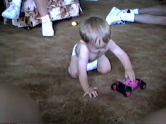 Vintage home video captures a curious baby crawling across the living room floor to play with a pink remote control toy car