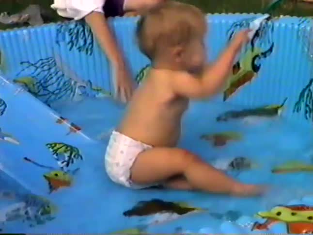 Vintage home movie footage captures a cute baby in a diaper splashing happily in a blue plastic kiddie pool