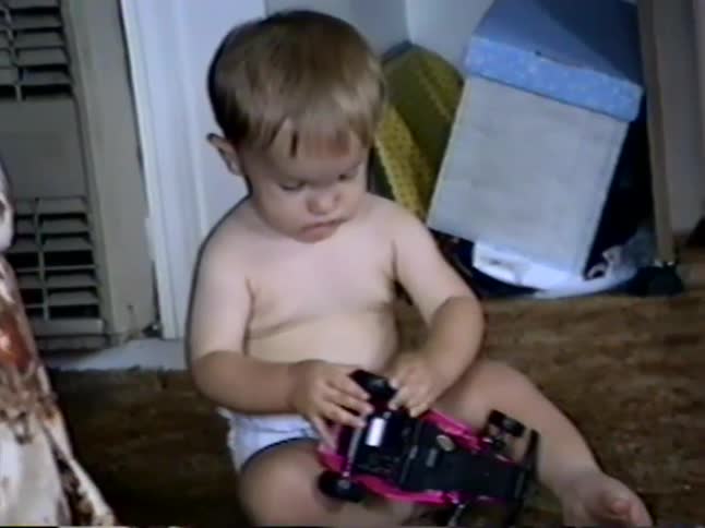 Vintage home video of a cute baby boy in a diaper playing with a pink toy car on the floor