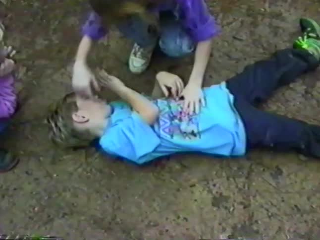 A young girl pretends to perform first aid on a laughing boy lying on the ground in a nostalgic home video from the 1990s