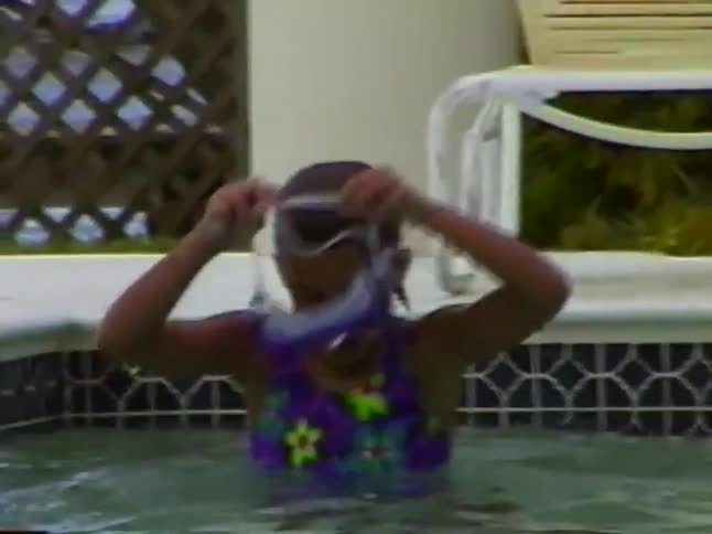 A young girl in a floral swimsuit excitedly puts on her swimming goggles in a pool
