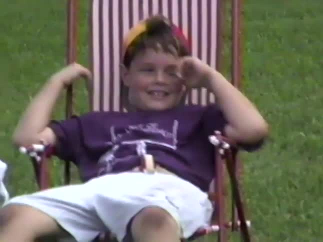 A young boy with a big smile sits in a lawn chair in the grass, laughing and being silly for the camera