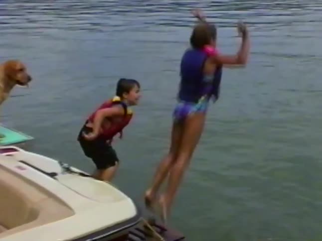 A boy and girl in life jackets playfully jump off the back of a boat into the water as their dogs watch in this vintage home video