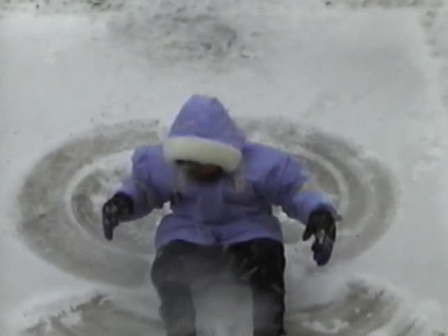 A high-angle vintage shot shows a child making a snow angel on a snowy patio before standing up to admire their work