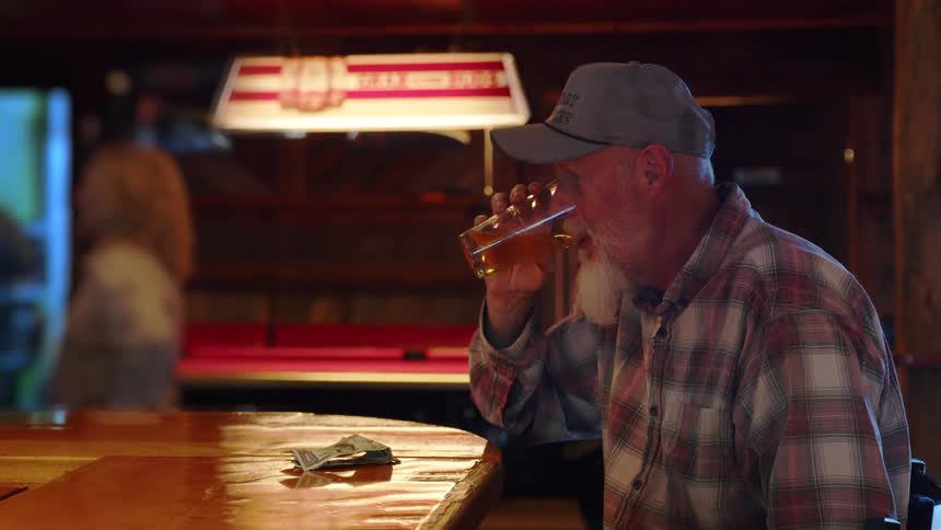 A senior man with a white beard sits alone at a rustic bar, pays for his drink, takes a sip of beer, and then walks away