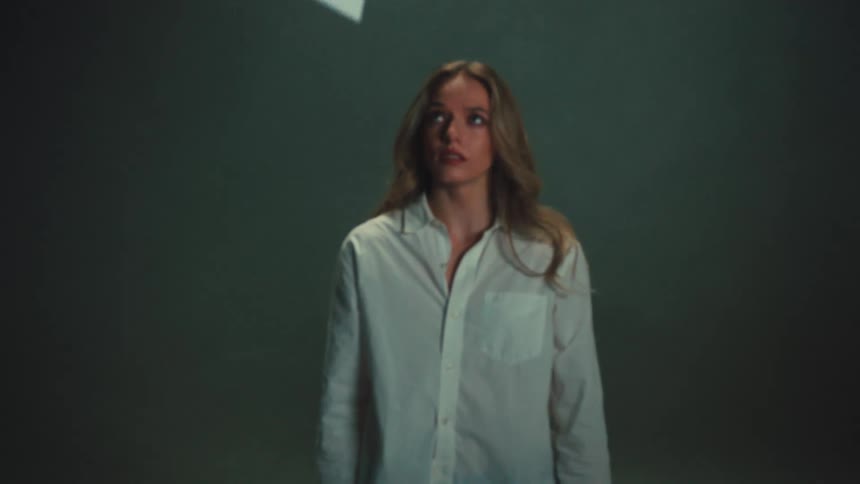 A fearful young woman stands still in a dark room as a white object slowly falls from above