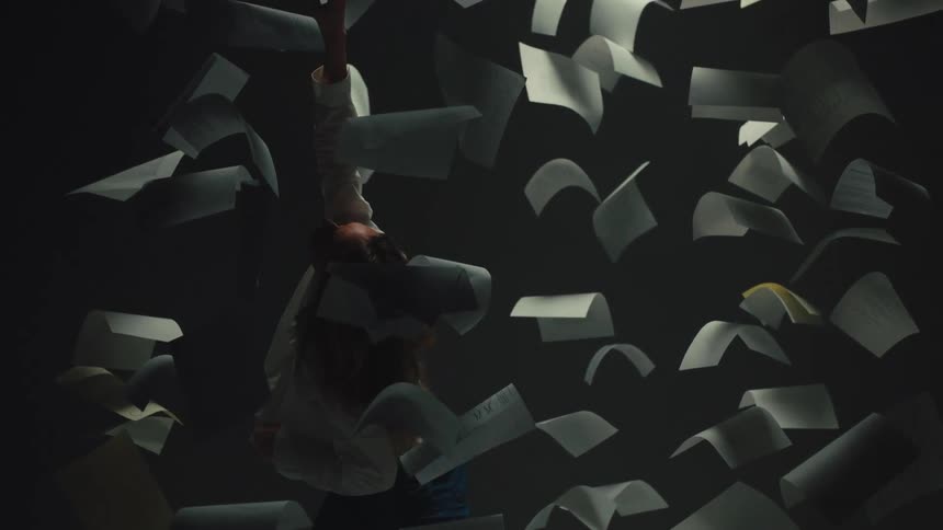 A woman in a white shirt and jeans floats in a surreal, dark space as sheets of paper fall around her in slow motion