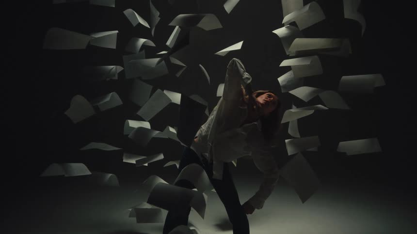 In a dramatically lit studio, a woman expresses anguish through contemporary dance while surrounded by a vortex of floating paper