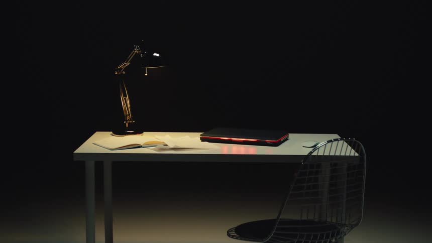 A minimalist desk setup with a laptop and lamp is illuminated by a single light source in a dark room