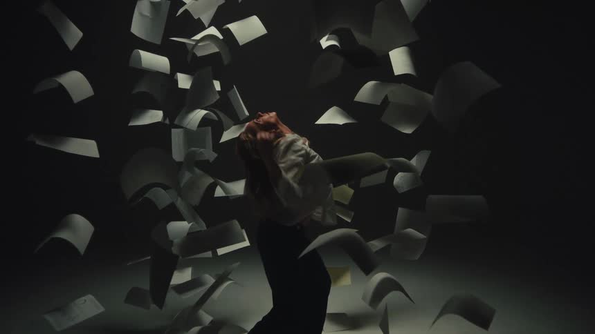 A woman collapses in slow motion as sheets of paper appear frozen in time around her in a dark, moody studio