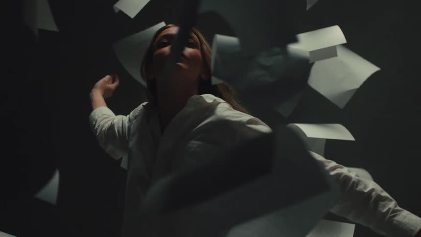 A woman gracefully floats through a chaotic storm of falling papers in a dark, atmospheric room in slow motion