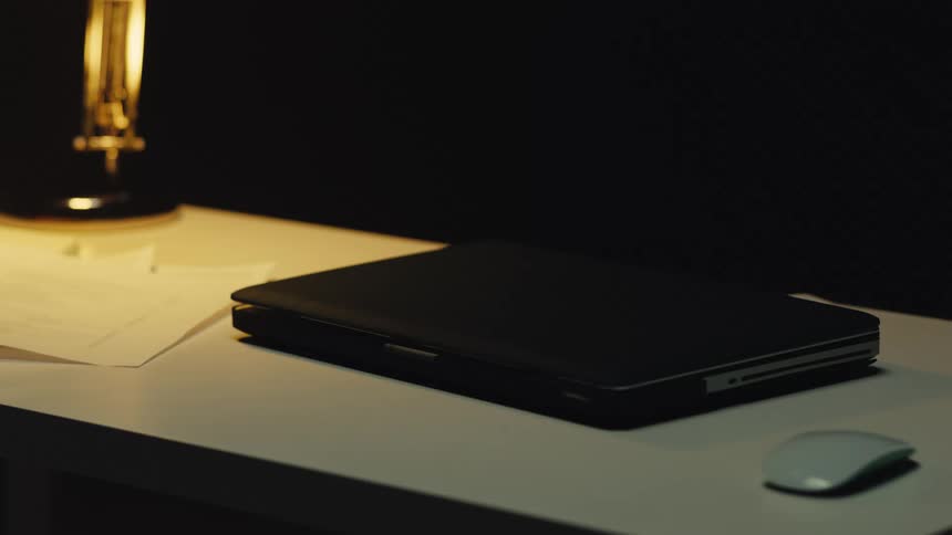 A closed laptop rests on a dimly lit desk next to a mouse and papers under the glow of a lamp