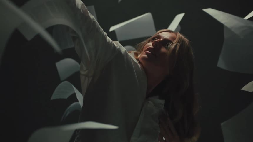 A woman in a dark room looks up as pieces of paper fly around her in slow motion