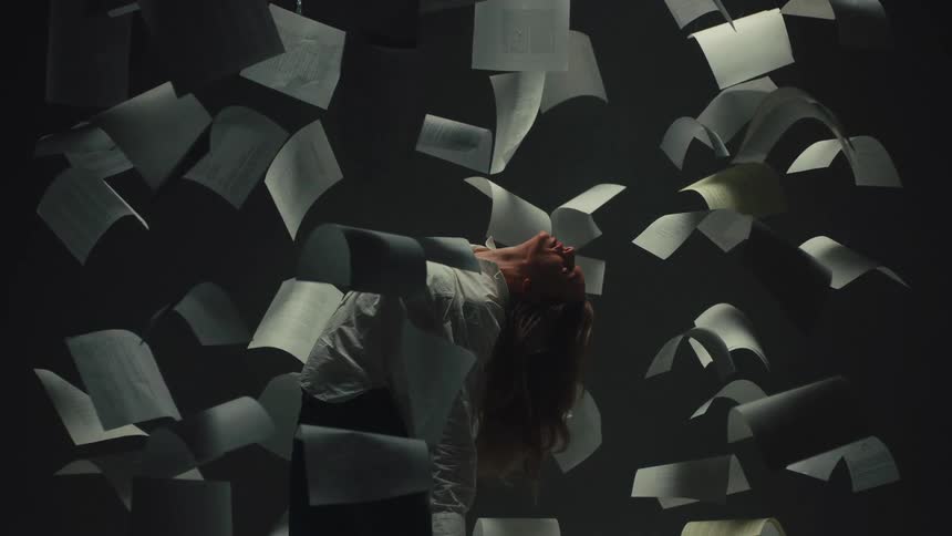 A woman is frozen in a dramatic pose amidst a cascade of suspended papers in a dimly lit room