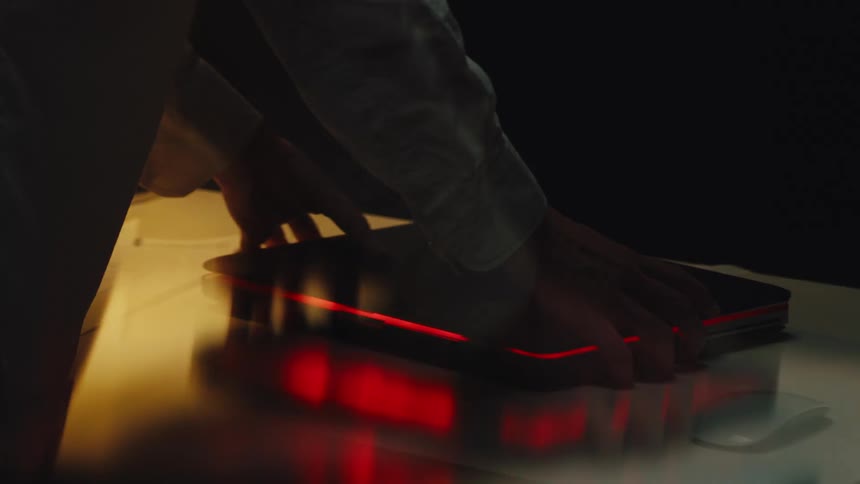 Close-up with motion blur of a person's hands frantically closing a laptop with a glowing red light in a dark room